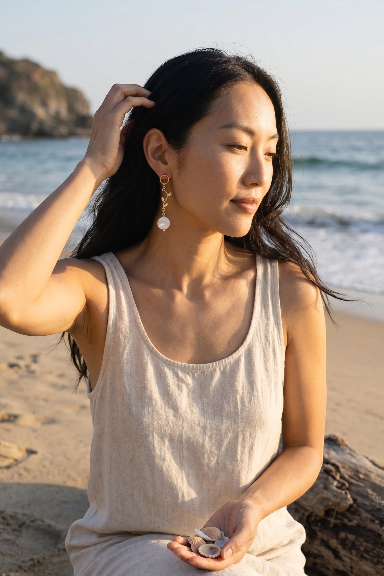 Sand and Sea 18k gold dangle earrings with white shell beads and leaf accents, worn by a woman on a beach.