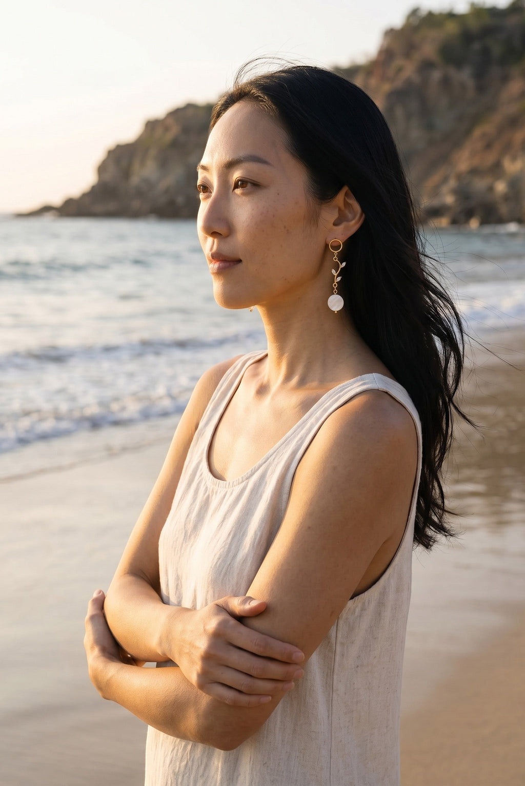Sand and Sea 18k gold dangle earrings with white shell beads and leaf accents, worn by a woman on a beach.