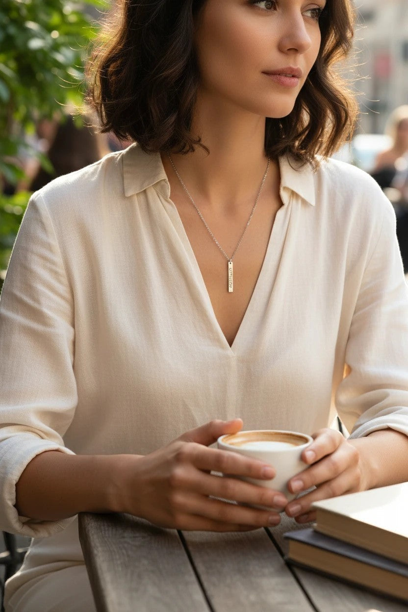 Sand and Sea engraved silver vertical bar necklace on a woman wearing a cream blouse, holding a coffee cup.
