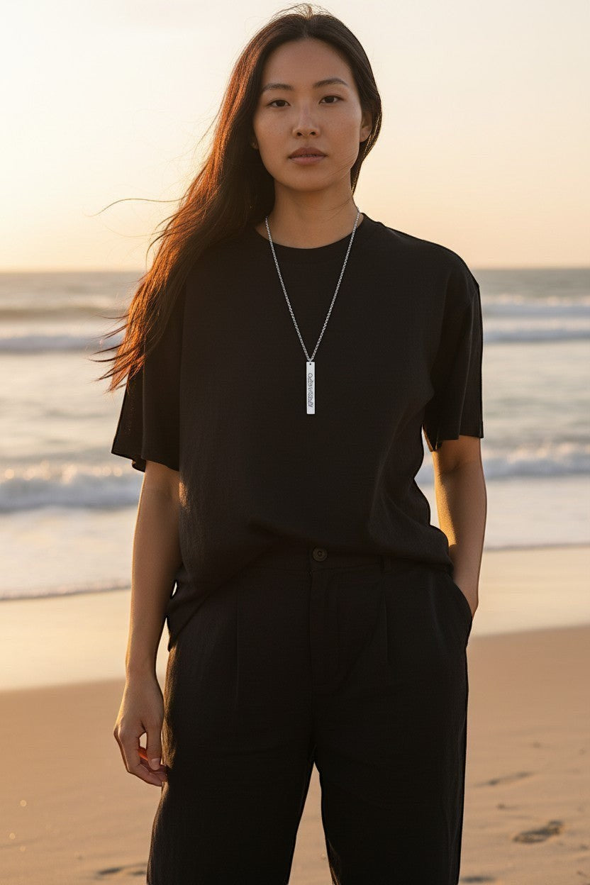 Sand and Sea engraved silver vertical bar necklace on a woman at the beach. Stainless steel pendant with laser engraving.