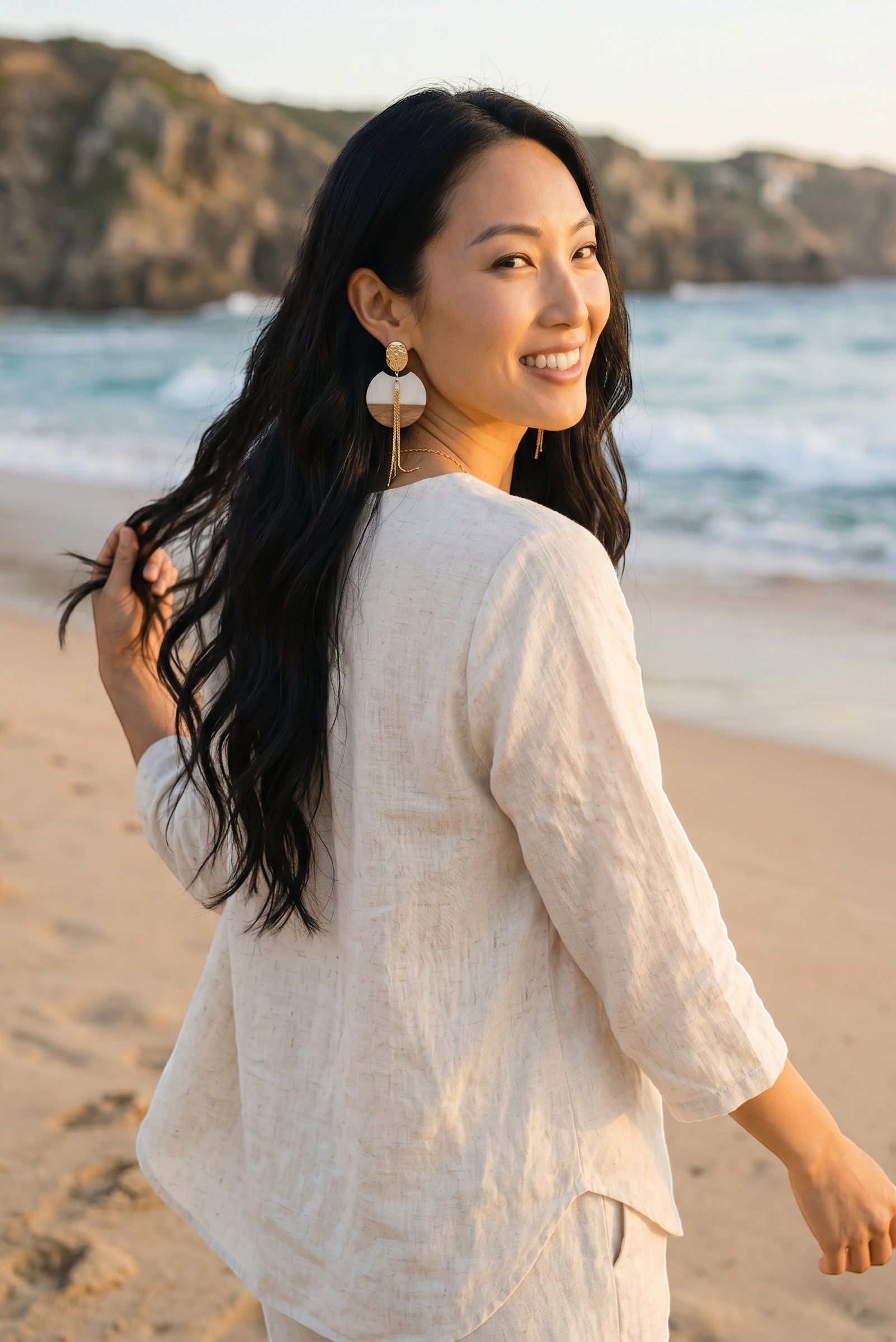 Sand and Sea geometric wood and gold metal drop earrings with cascading chains on a smiling woman at the beach.