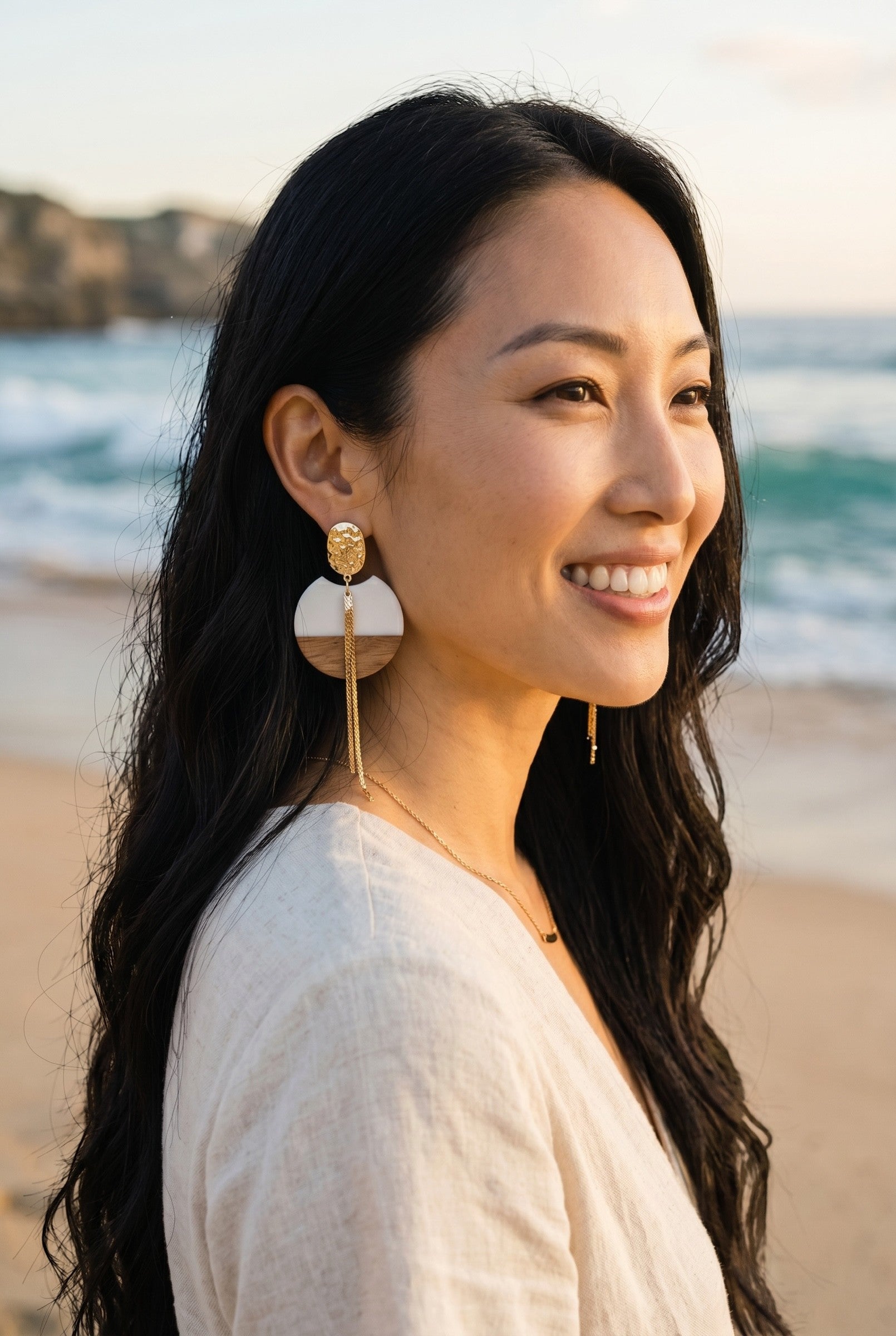 Sand and Sea geometric wood and metal drop earrings with gold chains. Model on beach.