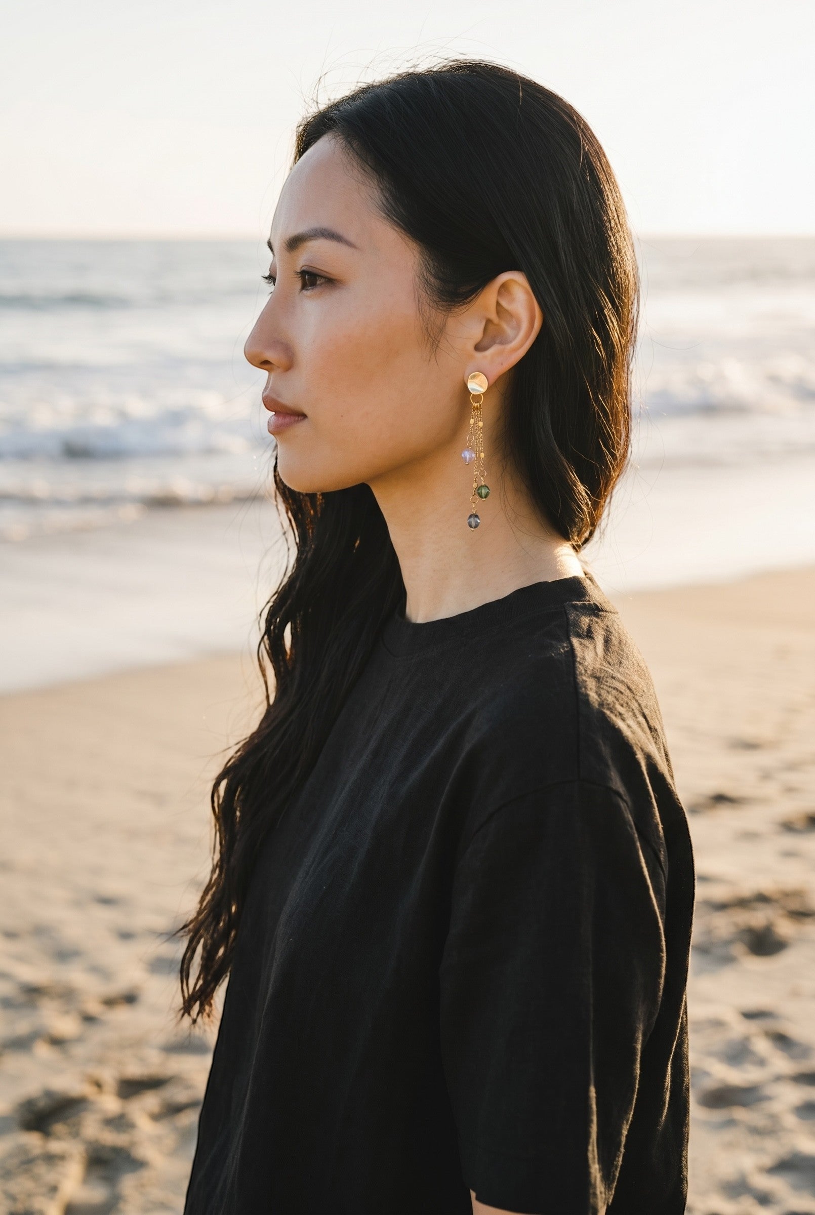 Sand and Sea gold plated drop earrings with pastel beads cascading on delicate chains. Model wears black shirt on beach.