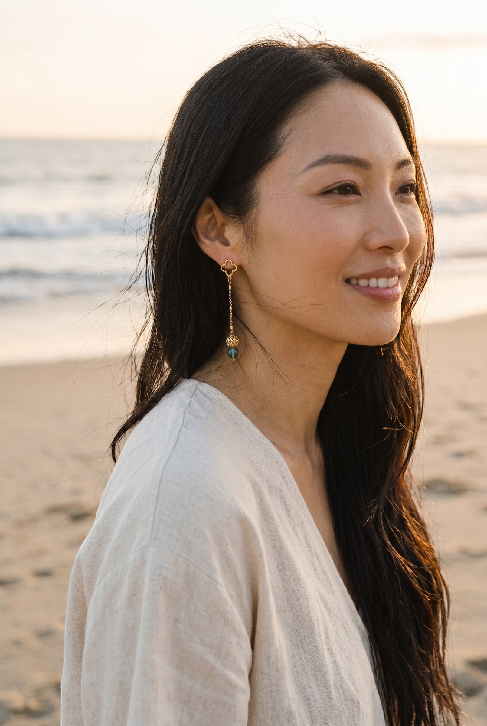 Sand and Sea gold-tone drop earrings with clover stud and blue gemstone bead. Model on beach.