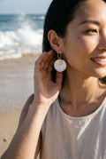 Sand and Sea white and cream marbled disc earrings with gold rectangular frame. Model on beach.