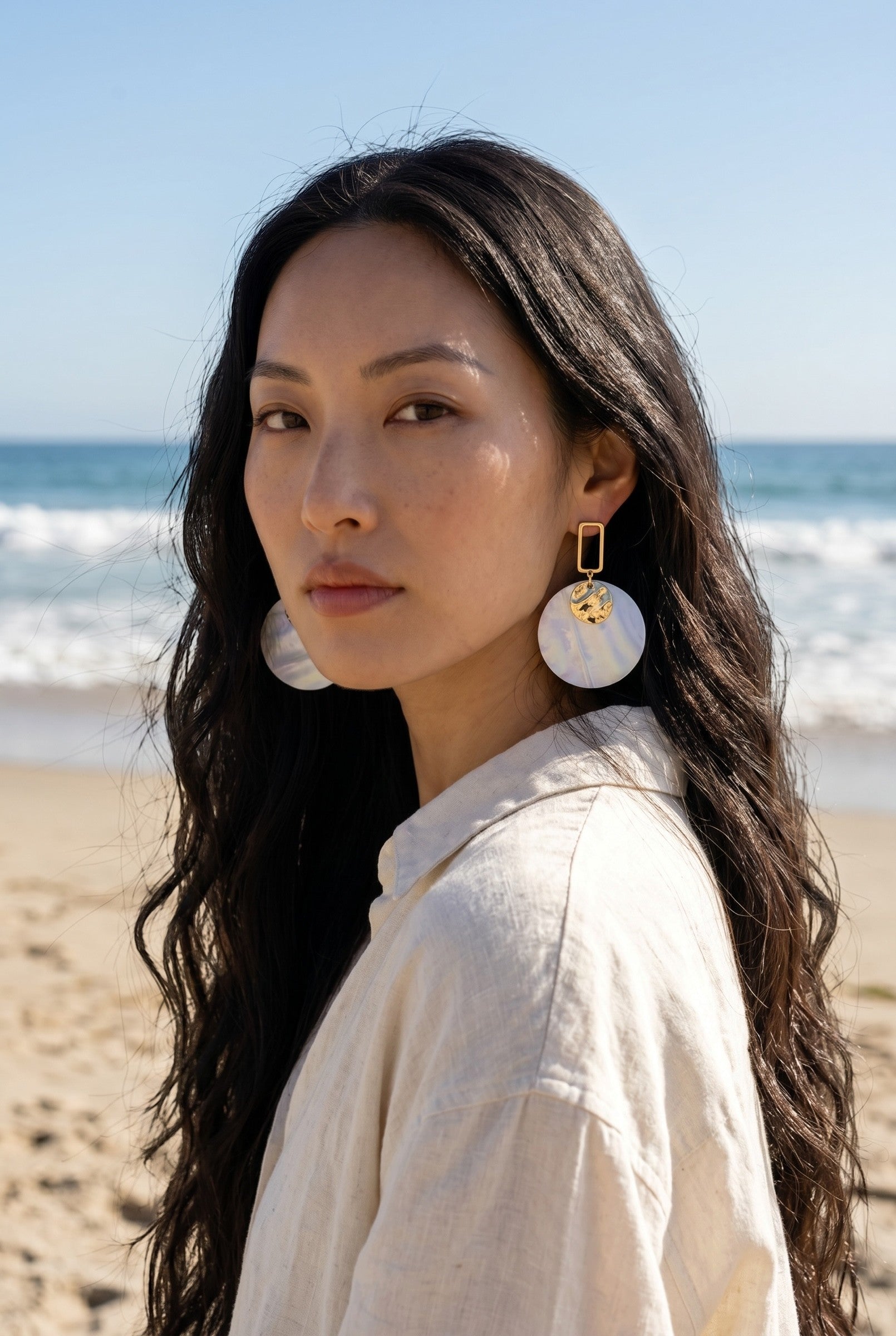 Sand and Sea white and cream marbled disc earrings with gold rectangular frame, worn by a woman on the beach.