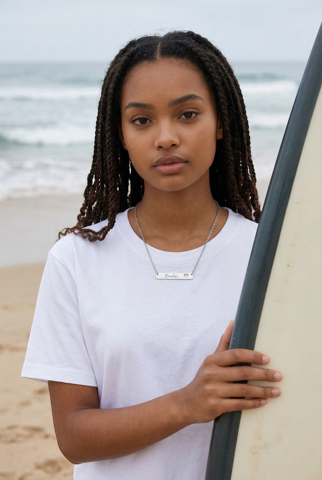 Sand and Sea silver bar necklace with cutout heart and "Borahae" engraving, worn by a young woman on a beach.