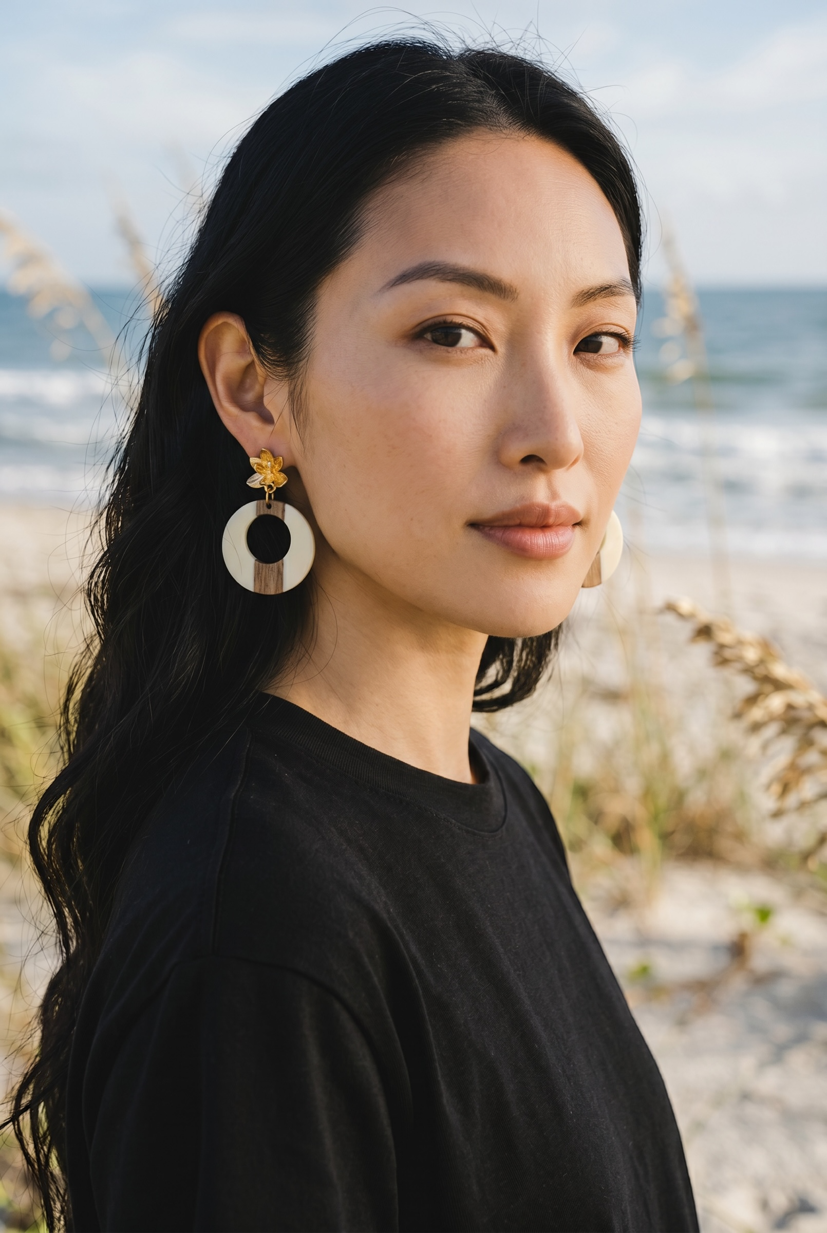 Sand and Sea statement hoop earrings with wood and cream accents and floral stud, worn by a woman on a beach.